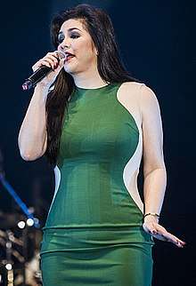 A long-haired middle-aged woman in a green fitted dress singing into a hand held microphone