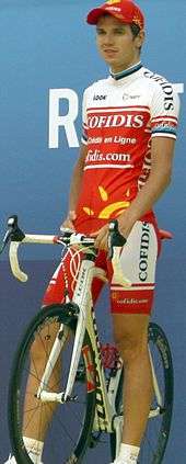 A man in his early twenties wearing a red and white cycling jersey with black trim, standing atop a stationary bicycle.