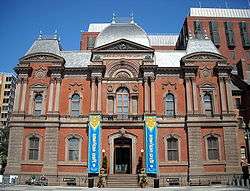 A 3-story, brick and red stone, second empire style building with a slate roof. The building is symmetrical with the entrance center and a blue and yellow banner on either side.