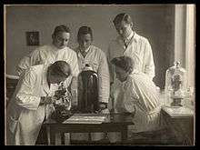 A woman in a white lab coat looks into a microscope while four other people in lab coats look on