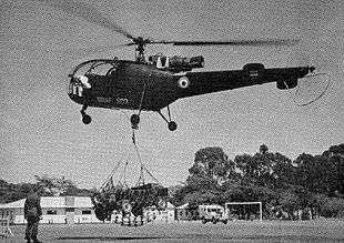 A green helicopter marked with a blue-white-red roundel; the roundel of the Federal Rhodesian Air Force. Against a backdrop of observers and barracks, it is lifting a Rhodesian Army jeep via a cargo net.