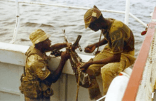 Two men in military fatigues handle a machine gun on the side of a boat