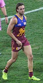 A man in a maroon, blue and gold football jersey and maroon shorts jogs on grass