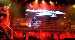 Singer on stage balcony flanked by musicians, below classic car on TV screen