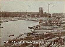 Sepia-tone photo of a pond surrounded by large logs. At the far end of the pond is a large building with a square tower and two smokestacks. Label is "T&T L CO MILL POND RICKETTS PA 1903" (i.e. Trexler and Turrell Lumber Company Mill Pond&nbsp;...)