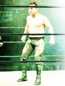 A color photo of a Mexican male wearing long black tights, standing in a wrestling ring