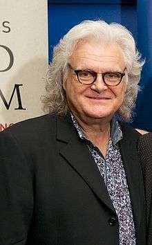 A man with long grey hair wearing glasses, a dark jacket and a blue and white shirt