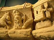 A photograph of a sandstone carving, broken into two pieces; on the left is the front half of a donkey, in the middle a fat man with a stick and a whip whilst on the right is a stylised windmill.