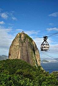 View of a peak situated near to the sea. A glass-paneled cable car passes suspended by cables.