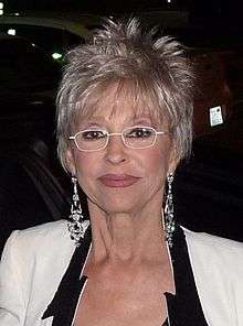 A woman is wearing glasses, a white dress, and earring is facing the camera.