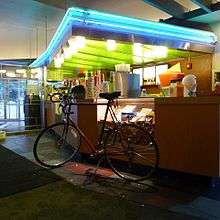 A bike sits parked before an indoor snack bar illuminated with blue neon lights.