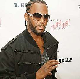 An image of R. Kelly standing on a red carpet and doing a hand sign towards the camera.