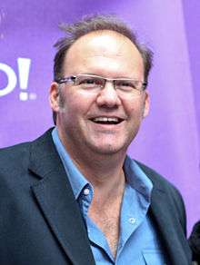 A smiling man with glasses wearing a blue shirt under a dark suit jacket.