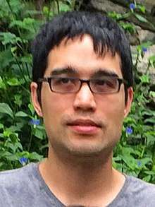 A man with short black hair and glasses seen from the neck up, where part of his gray T-shirt is visible. There are green leaves behind him