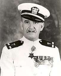 Head and shoulders of a white man with a thin, upturned mustache wearing a white peaked cap with black visor and a white jacket with dark shoulder boards, a row of medals on the left breast, and a star–shaped medal hanging from the neck.