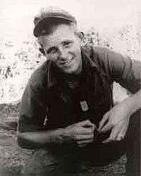 A smiling young man in military uniform, crouching with his forearms resting on his legs. His soft cap is pushed high up on his forehead and his dog tags hang out of his short-sleeved button shirt.