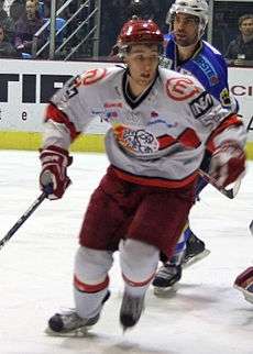 An ice hockey player skating towards the camera. He is wearing a red helmet and a red and white uniform.
