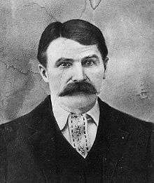 A head and shoulders shot of a middle aged white man with dark hair parted on his right and a full moustache, wearing a jacket and vest.