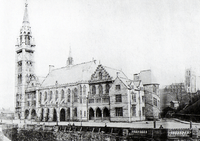 An aged greyscale photograph is filled with an elaborate building of craggy exterior and pointed spires is bathed in daylight. In the foreground is an empty road which drops sharply into a riverbank. In the background is a church.