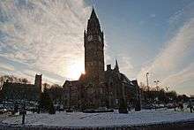 A fine and ornate building with a huge clock tower fills the central part of the image. It appears directly in front of the sun giving it a dark silhouette-like setting. The scene is marked by white snow.