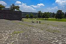 A large outdoor area with blackish-gray stone flooring of irregularly-cut medium-sized panels with mortar between them. Behind them in the distance are a grassy area with trees, buildings, and a blue sky with clouds