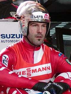 A man with a soul patch wears a red-and-white tight jumpsuit, with a red-and-white vest over it, and a metallic silver helmet with a raised full-faced visor. He is sat on the ground with his arms resting upon his legs.