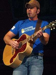 A man in a baseball cap, blue t-shirt and jeans, playing a guitar and singing into a microphone