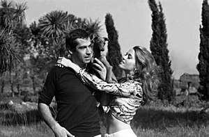 A man (Roger Vadim) standing with a dog on his shoulder with a woman (Jane Fonda) next to him.