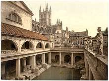 A late-nineteenth-century Photochrom of the Great Bath at the Roman Baths. Pillars tower over the water, and the spires of Bath Abbey&nbsp;– restored in the early sixteenth century&nbsp;– are visible in the background.