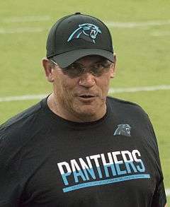 Color head-and-torso photograph of dark-haired Hispanic man (Ron Rivera), wearing a white and sky blue sport shirt and rectangular eyeglasses, seated at a press conference table with another man.