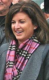 A photograph of a woman facing left and looking left while wearing a pink, black, and white plaid scarf, a grey jacket, and earrings