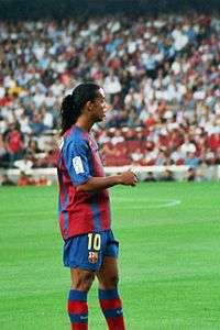Suntanned man with a long-haired hair, wearing a red and blue football shirt. In the background, green grass is rarely visible.