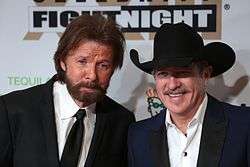 Two middle-aged men, one with brown hair and a beard, wearing a black jacket and necktie, the other with a grey mustache, wearing a black cowboy hat and dark blue jacket