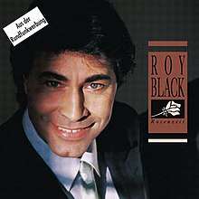 A closeup picture of a smiling man with black hair and blue eyes. He is wearing a black suit, a white shirt, and a white tie. On his right, there is an orange box with the words "Roy Black Rosenzeit" and a stylised white drawing of a rose.