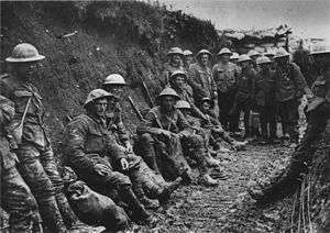 In a black and white photograph, 15 soldiers in standard uniform are leaning against a trench wall
