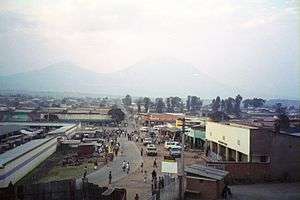 Photograph of Ruhengeri, Rwanda, with buildings, a street, and people visible, and mountains in the background, partially in cloud
