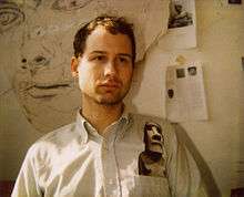 Young man in an art studio, in front of a large drawing and art clippings taped to the wall.