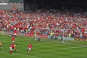 An action scene from a soccer match, played on a sun-soaked summer's day. Before an old-fashioned terraced stand packed to the rafters with fans, mostly clad in red, a penalty kick has just been taken by a player wearing a red shirt and white shorts. The ball is nestled in the bottom-right-hand corner of the net, with the goalkeeper helpless on the opposite side of his goal. Behind the penalty taker, a few players from each team can be seen on the edge of the penalty area.