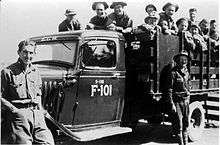 Black and white image of a stakebed truck from the 1930s with two men standing in front and about a dozen men in the back. The truck is labeled ECW (Emergency Conservation Work, original name of the CCC) above the windshield, and "S-118 F-101" on the door.