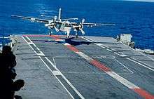 A large propeller-driven aircraft is moments away from landing on the flight deck of an aircraft carrier