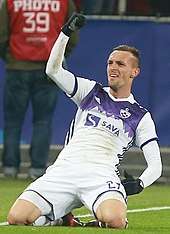 A short-haired white man in a white-purple football kit, sitting on his knees with a raised fist, celebrating a goal.