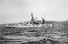 A large light grey battleship tilts back in choppy water as it slowly sinks.