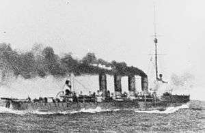 Waves crash against the bow of a cruiser as it steams ahead with large clouds of smoke coming out of all four of its funnels.