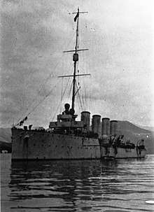 A cruiser sits motionless in the water, with four large funnels, a bridge, and a main mast prominently appearing aboard the ship. A small board lies next to the ship in the foreground, while hills lie in the background.