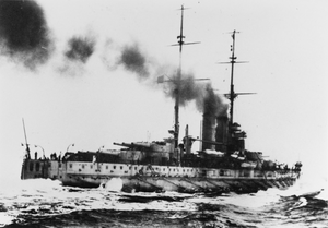 A large battleship steaming away through the water at high speed. Its stern can be seen in the foreground with water hitting the sides of the ship. Smoke can be seen billowing out of the funnels of the ship.