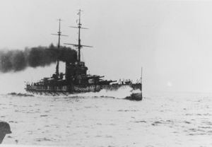 A large battleship steams through the water. Water breaks against the bow as heavy dark smoke emerges from the ship's two funnels.