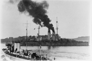 A large battleship steams through a harbor. Large clouds of smoke can be seen coming from the ship's funnels while a smaller vessel is sailing in the foreground. Hills and the coastline can be seen in the background.