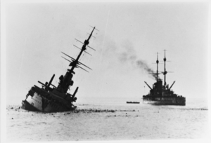A battleship lies low in the water with a heavy list after being struck by a torpedo. Another battleship can be seen floating in the background close by.