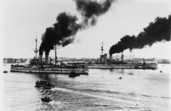 Two battleships steam into a harbor with several smaller boats around them. The larger battleship is in the background while the smaller one is in the foreground.