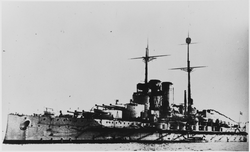 A large battleship sits at anchor. A small boat can be seen moored next to the battleship in the foreground.